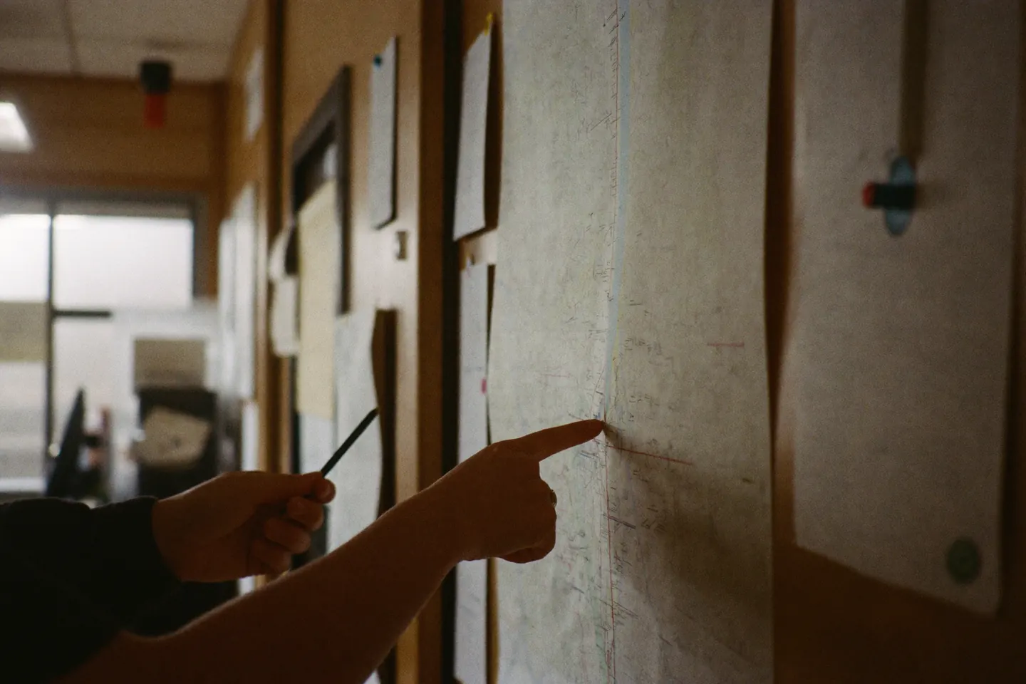 Two hands at a wall of pinned paper notices in a small agency office, one pointing at a document while the other holds a pen.